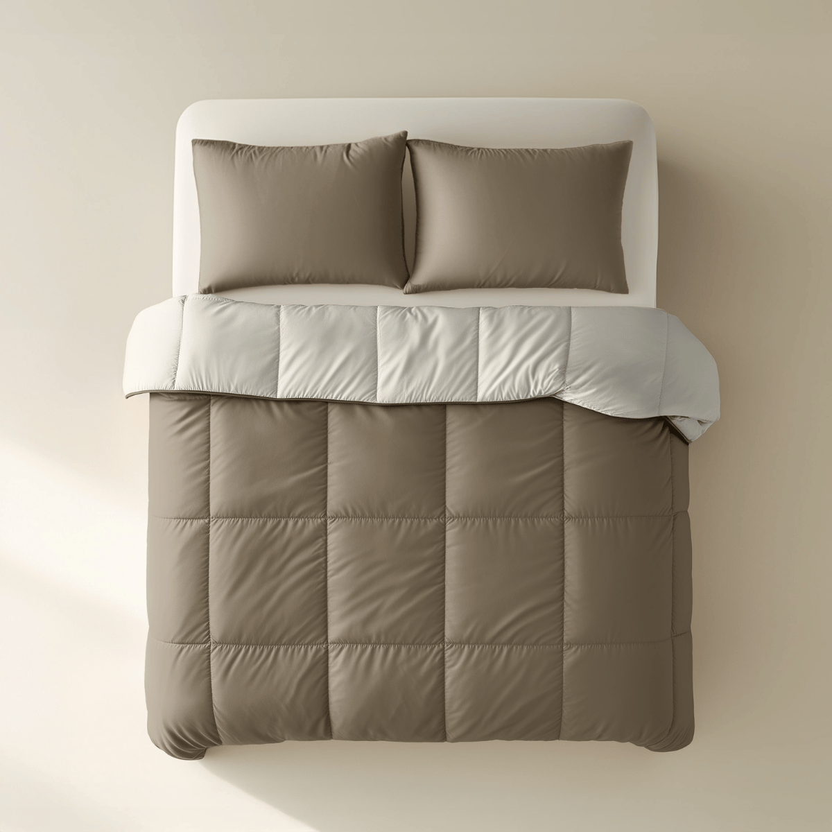 Ein gemachtes Bett mit zwei braunen Kissen und einer taupefarbenen Bettdecke aus dem Zelesta Premiumbed Baumwolle - Taupe & Cream, bedeckt mit einer gefalteten hellgrauen Decke, von oben gesehen vor einem beigen Hintergrund.