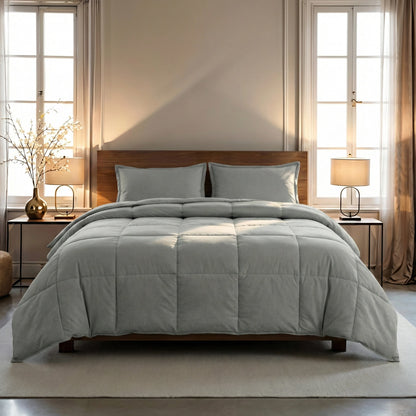 Ein ordentlich gemachtes Bett mit dem Zelesta Wonderbed 4 Seasons Double Baumwolle in Morning Grey und zwei Kissen, flankiert von Nachttischen mit Lampen, in einem sanft beleuchteten Schlafzimmer mit großen Fenstern und neutralem Dekor.