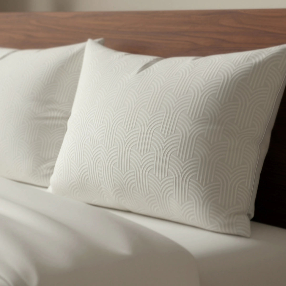 Zwei cremefarbene Zelesta Flanel Art Kissenbezüge (Set von 2, 80x80cm) mit dezentem geometrischen Muster liegen auf einem ordentlich gemachten Bett mit weißen Laken und lehnen an einem hölzernen Kopfteil.