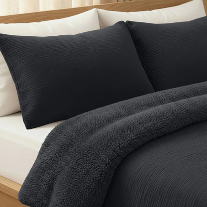 Ein ordentlich gemachtes Bett mit zwei Teddy Baumwolle Mousseline Kissenbezügen Set von 2 - Anthrazit, einer strukturierten schwarzen Decke und einem schwarzen Plüschüberwurf auf einem hölzernen Bettrahmen.