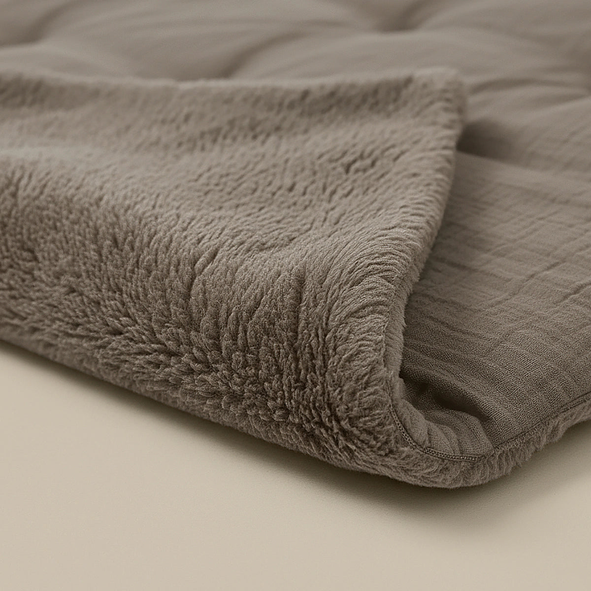 Eine Nahaufnahme des Zelesta Teddy Baumwolle Musselin - Taupe zeigt seine weiche, gefaltete Textur mit einer Plüschseite und einer glatten, gesteppten Seite auf einer neutralen Oberfläche - perfekt für gemütliche Momente und temperaturregulierenden Komfort.