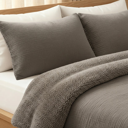 Ein ordentlich gemachtes Bett mit taupefarbenen Teddy Baumwolle Mousseline Kissenbezügen Set von 2 und einer passenden Bettdecke, gekrönt mit einer plüschigen grauen Sherpa-Decke, vor einem hölzernen Kopfteil - perfekt für gemütlichen Komfort.