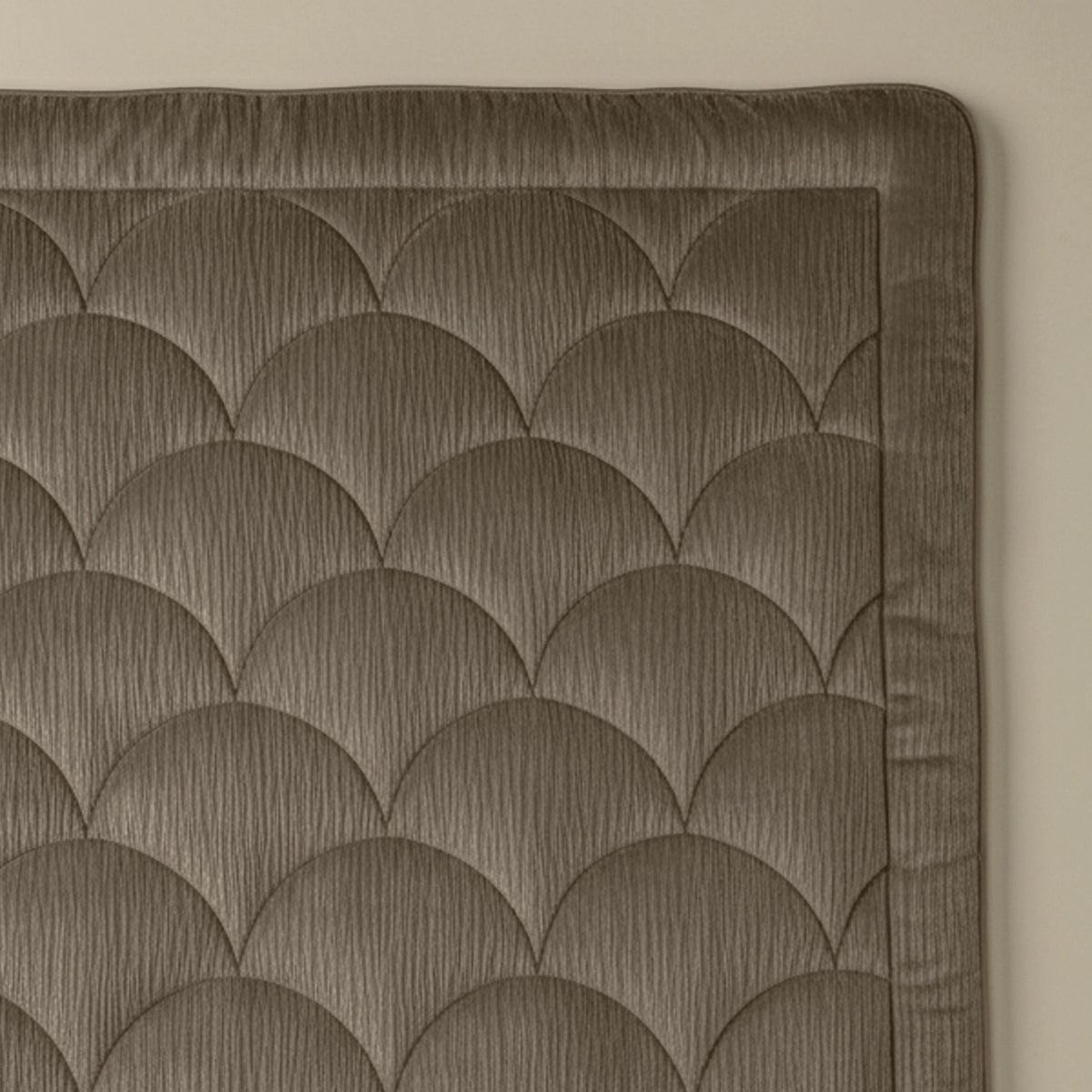 Nahaufnahme des Zelesta Flanel Velvetbed Chique - Taupe mit gestepptem Wellenmuster in gedämpftem Braun und unifarbener Umrandung vor einer hellbeigen Wand.
