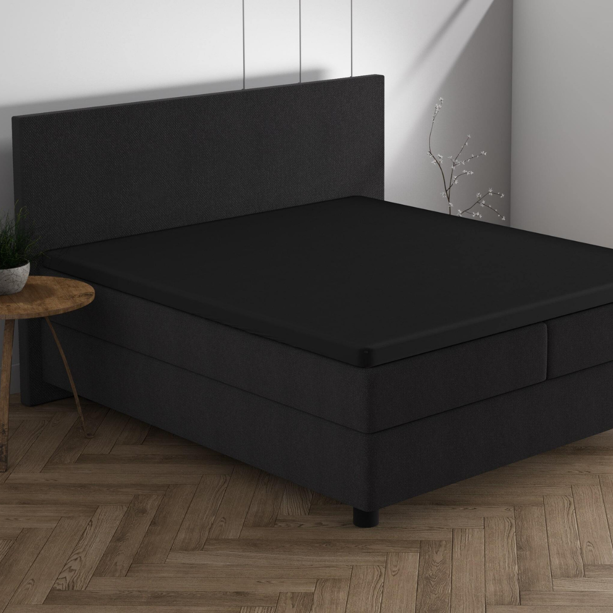 Ein moderner Dreamhouse-Rückzugsort: ein schwarzes Polsterbett mit dem Doppel Jersey Topper Spannbettlaken - Schwarz, auf einem Fischgrät-Holzboden, daneben ein runder Beistelltisch mit einer Pflanze.
