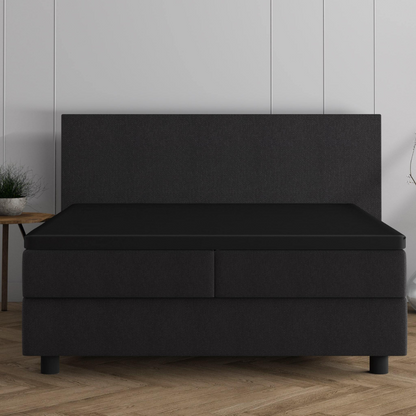 Ein modernes, dunkles Bett mit hohem Kopfteil und zwei Schubladen, bekleidet mit dem Doppel Jersey Topper Spannbettlaken - Schwarz, steht vor einer weiß getäfelten Wand auf einem Holzboden.